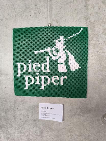 A green rectangle made out of Hama beads. In white beads there are the words "pied piper" and the Pied Piper of Hamelin. It's a little guy with a cape and a little hat, holding a pipe.