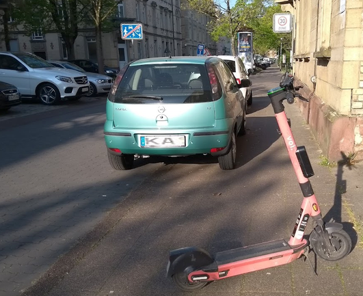 Scooter von voi steht quer über Gehweg, vor halb auf Gehweg parkendem Auto und zwingt Fußgänger zum Slalom.