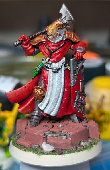 A sturdy figure in armour and long red robes. She has a large warhammer slung over one shoulder and holds a shield in her other hand. Her head is bare and her hair has been shaved close at the sides