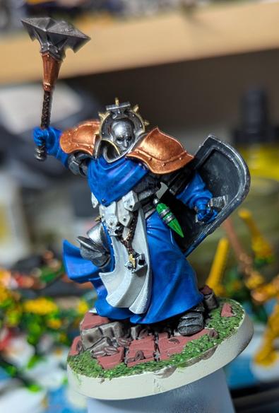 A sturdy figure in armour and long blue robes. He holds a large warhammer aloft and has a shield in his other hand. His helmet has a spiked halo reminiscent of a Catholic saint, and a beaded belt at his waist bears an icon of an anvil and lightning bolt