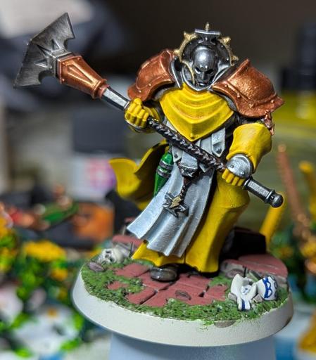 A sturdy figure in armour and long yellow robes. He wields an enormous two-handed warhammer with no shield, and wears the same helmet as his blue colleague. The base for all three models suggests terracotta tiles overgrown with moss