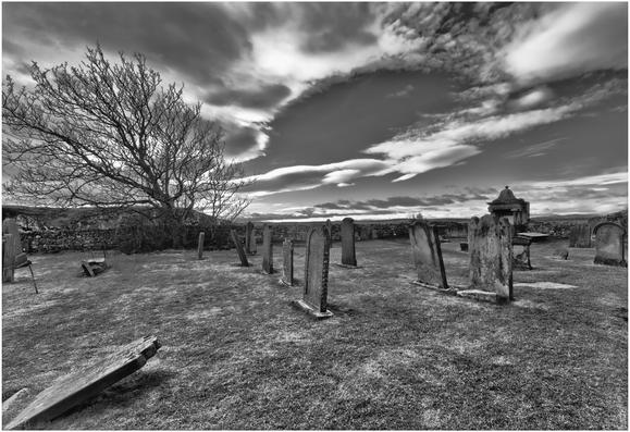 A black and white image of a cemetery featuring several gravestones of varying sizes. In the background, there is a bare tree and a stone wall under a dramatic sky with clouds. The ground is grassy and some gravestones are leaning or partially toppled.