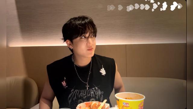Changbin during a livestream from his hotel room. He's sitting on a couch and has some mostly empty foot containers in front of him. He's wearing a skz dwaekki sleeveless shirt with a couple of dwaekki charms pinned to it, and a cross pendant on a chain. He's looking off screen.