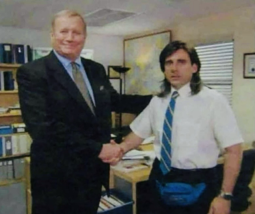Zwei Männer stehen in einem Büro und schütteln sich die Hand. Der Mann links trägt einen dunklen Anzug und lächelt, während der Mann rechts ein weißes kurzärmliges Hemd, eine blaue Krawatte und eine auffällige blaue Bauchtasche trägt. Die Szene wirkt wie aus den späten 90er- oder frühen 2000er-Jahren.