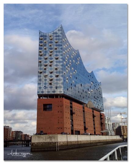 Farbfoto im Hochformat. Es zeigt eine Außenansicht der Elbphilharmonie in Hamburg unter einem leicht bewölkten Himmel. Das Gebäude ist das zentrale Element des Bildes und nimmt fast den gesamten Bildausschnitt ein. 
Es besteht aus zwei deutlich unterscheidbaren Teilen: einem modernen, wellenförmigen Oberbau aus Glas und einem unterirdischen, roten Backsteinteil. Der gläserne Oberbau ist mit vielen kleinen, runden Fenstern versehen, die sich in einem regelmäßigen Muster befinden und dem Gebäude ein einzigartiges Aussehen verleihen. Die Fenster reflektieren den Himmel und die Umgebung, wodurch sich das Gebäude dynamisch in das Umfeld einfügt. Der untere Teil des Gebäudes aus roten Backsteinen wirkt im Vergleich zum oberen Teil massiver und eher traditionell. Er ist mit regelmäßig angeordneten, kleinen Öffnungen versehen. 
Das Gebäude steht direkt am Wasser, und man sieht einen Teil einer Betonmauer und darauf einige parkenden Autos. Im Hintergrund sind weitere Gebäude der Hamburger HafenCity zu sehen, sowie einige Kräne.

(Text mit Hilfe von KI generiert)