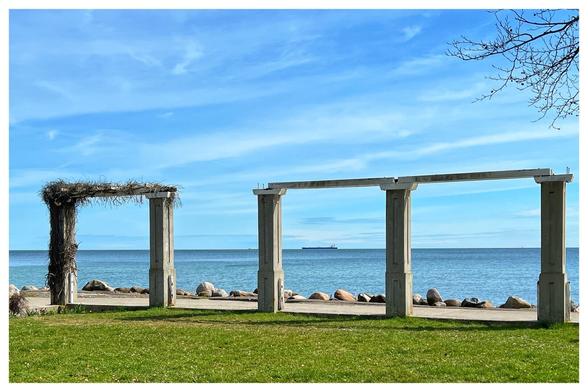 Blick durch zwei eckige Arkaden auf die blaue Ostsee unter strahlend blauem Himmel mit wenigen Schleierwolken. Im Hintergrund ein Schiff auf Reede