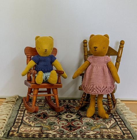 Maileg Teddy mum, in a pink cotton dress. and Teddy junior in checked cotton shorts and a knitted blue tee shirt are sitting in rocking chairs.