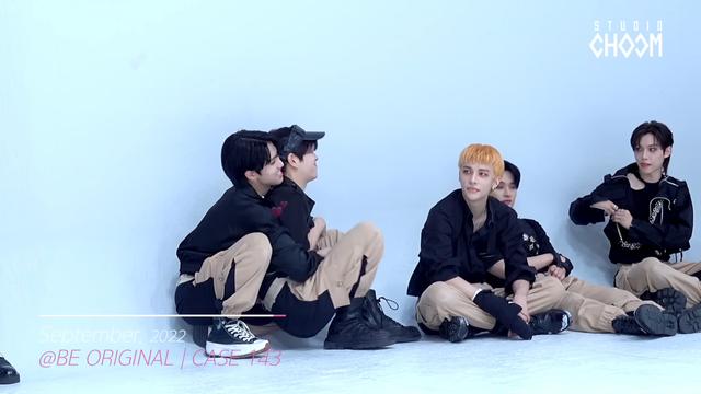 Seungmin crouching down but off-balance because Han is crouching behind him and has both arms around him, pinning Seungmin's arms and destabilising his centre of gravity. Han is pleased about this.