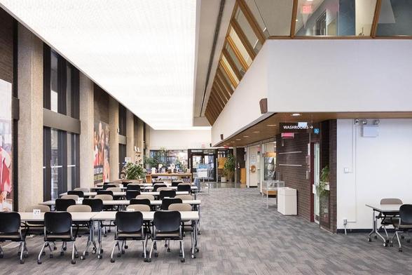 Interior of a Brutailist library, an L-shaped reading room with a row of windows framed by concrete columns to the left. To the right is a two-level structure with slanted windows. Rows of tables and chairs fill the space.