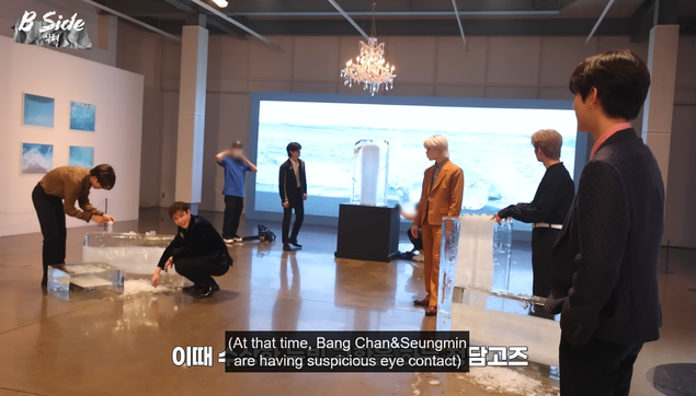 In an art gallery, Bang Chan is crouched near where Han is making a tower out of ice fragments. He's making eye contact with Seungmin across the room. Caption: (At that time, Bang Chan & Seungmin are having suspicious eye contact)