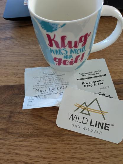Ein Photo. Karten vom Bergwipfelpfad und der Skywalk Hängebrücke in Bad Wildbad; dahinter eine weiße Tasse: 'klug wars nicht, aber geil!'