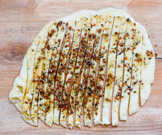 ein Stück ausgerollter Teig, mit Gewürzöl bestrichen undlängs in Streifen geschnitten, oben ist der Teig nicht durchgeschnitten und verbindet die einzelnen Streifen.