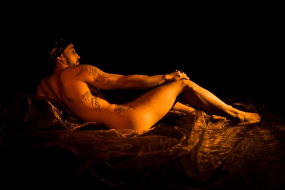 The image features a person lying down on a fabric with artistic and dramatic lighting. The person appears to be unclothed but tastefully posed, with tattoos visible on their body. The setting and lighting give the image an artistic or classical feel.