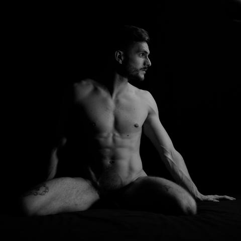 The image is a black and white photograph of a nude man sitting with one leg bent and the other extended to the side. He is looking away from the camera. The lighting emphasizes his muscular physique against a dark background.