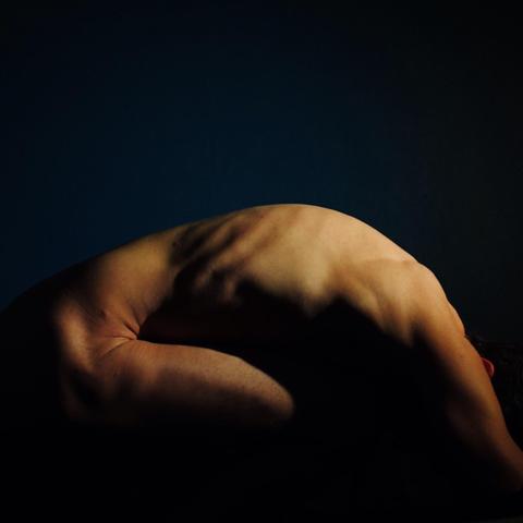 The image is an artistic photograph of a person in a curled, fetal-like position. The focus is on the contours and shadows of their back and arms, creating a dramatic effect against a dark background.