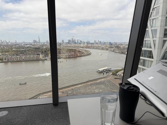 View from a high rise in Canary Wharf onto the Thames river, showing its brown water and a few ferry boat passing by. It’s a sunny day. In the distance you can see the large buildings of central London.