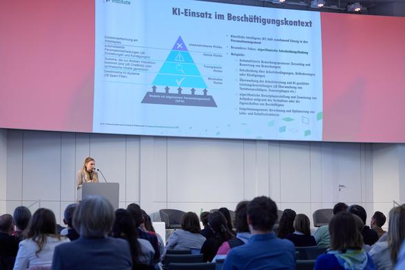 Das Bild zeigt Madeleine Müller, die vor einer großen Zuhörerschaft über den Einsatz von künstlicher Intelligenz im Beschäftigungskontext spricht, wobei ein Schaubild auf der Leinwand eine Pyramide mit verschiedenen Risikostufen der KI-Anwendungen darstellt. Die Pyramide zeigt Modelle mit allgemeinem Verwendungszweck (GPAI) an der Basis, mit steigenden Risiken nach oben hin: minimales, Transparenz-, hohes und unannehmbares Risiko. Die einzelnen Risikostufen werden mit Beispielen für KI Anwendungen im Personalwesen erläutert, wie z.B. Bewerbungsprozesse, Leistungsbeurteilungen und Entgeltmanagement. Im Vordergrund sitzen sehr viele Zuschauer*innen.