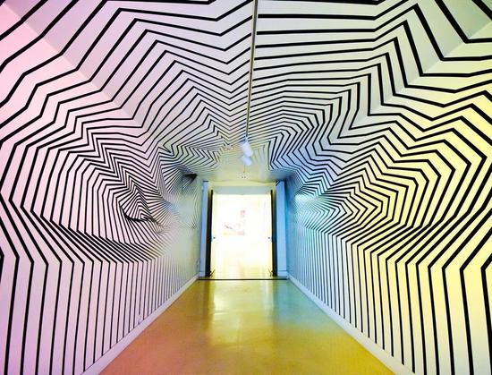 A long white-walled hallway covered in black tape lines creating jagged and curving indentations across the walls and ceiling, with a yellow-lit floor leading to an open doorway emitting yellow light.