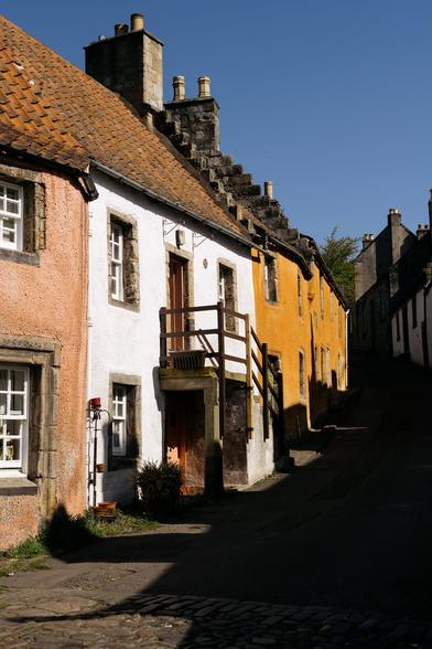 A row of brightly painted historic cottages in shades of pink, white, and mustard yellow line a cobbled village street. The buildings feature small windows, wooden doors, and stepped gables, all under a vivid blue sky.