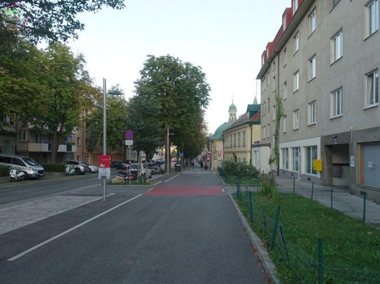 Städtische Straße mit gemeinsamem Geh- und Radweg, Bäumen, Wiese, im Hintergrund eine Kapelle