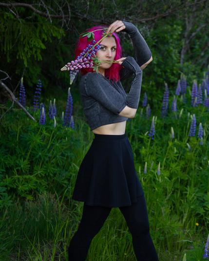 A pink haired woman in nature holding two lupine flowers in front of her face