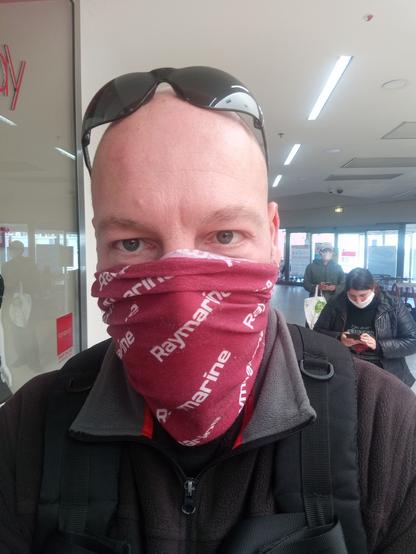 Selfie of a bald guy wearing dark sunglasse on the top of his head and a Raymarine scarf over nose and mouth. He's standing in a queue in a minimally lit hallway outside a supermarket, waiting his turn to be allowed to buy food.