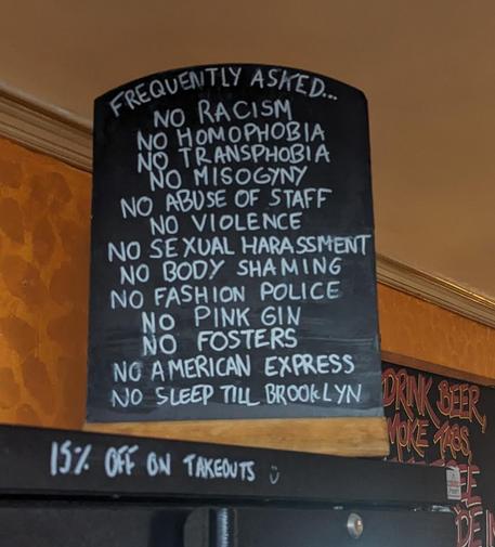 A chalk board that reads:
NO RACISM 
NO HOMOPHOBIA
NO TRANSPHOBIA
NO MISOGYNY
NO ABUSE OF STAFF
NO VIOLENCE
NO SEXUAL HARASSMENT 
NO BODY SHAMING
NO FASHION POLICE 
NO PINK GIN 
NO FOSTERS
NO AMERICAN EXPRESS 
NO SLEEP TILL BROOKLYN