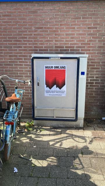 Leeg reclame frame op elektriciteitskastje met poster: Bovenaan staat in grote zwarte hoofdletters “HUUR OMLAAG”, daaronder, op de achtergrond een graphic van twee omgedraaide, rode rijtjeshuizen, de punten van de daken wijzen omlaag. Op de voorgrond staat een tekst in witte letters: Landelijke demonstratie in Utrecht, Zaterdag 10 mei, 14.00, Domplein, woonprotestutrecht.nl 
Blauwe fiets met bruin zadel ernaast geparkeerd. Bakstenen muur.