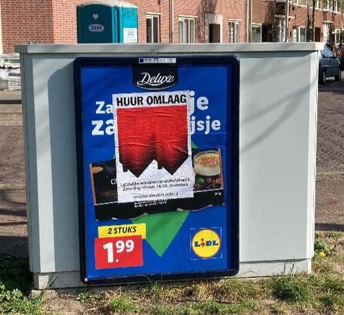poster over reclemae bord op kastje: Bovenaan staat in grote zwarte hoofdletters “HUUR OMLAAG”, daaronder, op de achtergrond een graphic van twee omgedraaide, rode rijtjeshuizen, de punten van de daken wijzen omlaag. Op de voorgrond staat een tekst in witte letters: Landelijke demonstratie in Utrecht, Zaterdag 10 mei, 14.00, Domplein, woonprotestutrecht.nl
Op de achtergrond woningen en een dixie