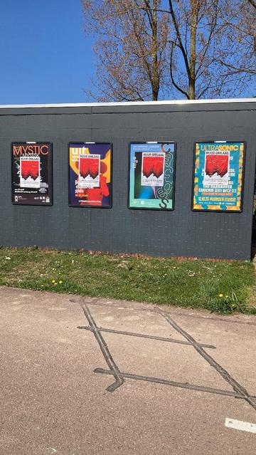 posters geplakt op 4 reclame borden op huisje met donker geverfde bakstenen, Bovenaan staat in grote zwarte hoofdletters “HUUR OMLAAG”, daaronder, op de achtergrond een graphic van twee omgedraaide, rode rijtjeshuizen, de punten van de daken wijzen omlaag. Op de voorgrond staat een tekst in witte letters: Landelijke demonstratie in Utrecht, Zaterdag 10 mei, 14.00, Domplein, woonprotestutrecht.nl
Boom achter en blauwe lucht, voor gras en fietspad