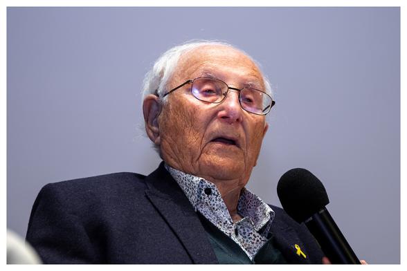 KZ-Überlebender Albrecht Weinberg als querformat Portraifoto. 
Ein hundert Jahre alter Mann mit Falten, Altersflecken, Brille und weißem schütteren Haar spricht in ein von einem anderen Menschen gehaltenes Mikrofon.
Um Das Bild ist ein kleiner weißer Rahmen.