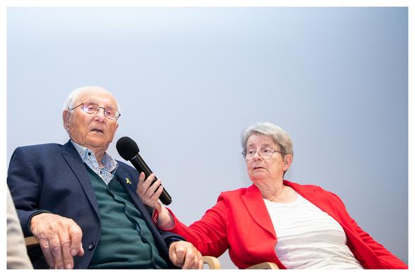 KZ-Überlebender Albrecht Weinberg linksseitig im Bild. Spricht in das Mikrofon, welches von seiner Mitbewohnerin und Freundin Gerda Dänekas gehalten wird. 
Um das Bild ist ein kleiner weißer Rahmen.