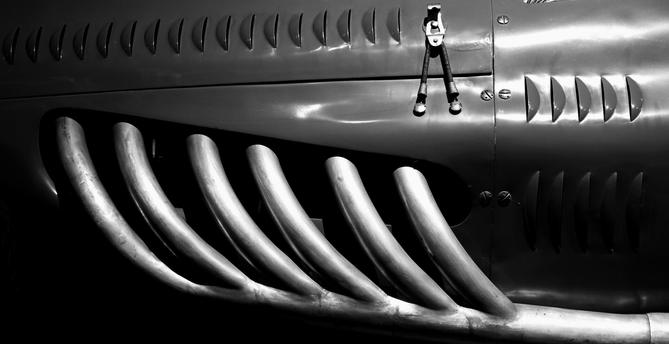 close-up of a vintage Bugatti's metallic bodywork, showcasing a series of prominent, curved exhaust pipes. The design highlights sleek, aerodynamic lines and textured details, all rendered in black and white for a dramatic effect. The interplay of light and shadow emphasizes the car's classic features.