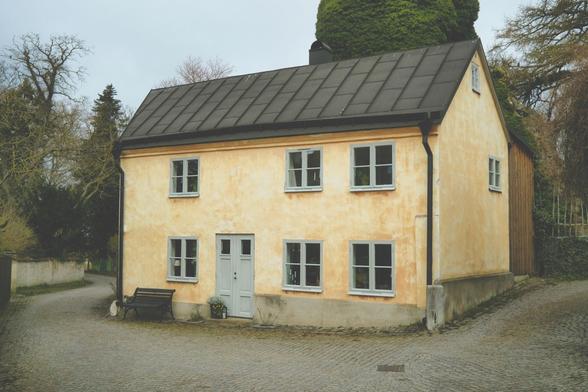 Pittoreskt hus i sten, blekgult till färgen och byggt i klassiskt gotländsk stil, mitt inne i Visby. I bakgrunden skymtar toppen av Sankt Olofs Kapell, övervuxen av murgröna.