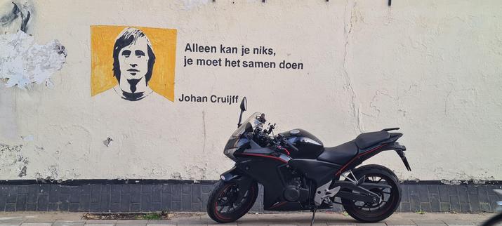 A black with red pin striping Honda motorcycle parked on the sidewalk in front of a somewhat damaged cream white stuccoed wall with a black brick band at the bottom. 

On the wall, above and to the left of the bike a black and white portait of a young Johan Cruijff, a narrow faced man with shoulder lenght hair, in a yellow square next to the text Alleen kan je niks. Je moet het samen doen. Which is Dutch for Alone you can accomplish nothing. You need to do it together.
