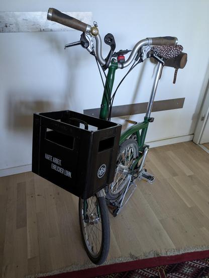 Ein grünes Brompton Faltrad. Vorn auf dem Frontcarrier ein leerer Kasten Bier der Brauerei Bergmann aus Dortmund mit dem Claim: Harte Arbeit. Ehrlicher Lohn.