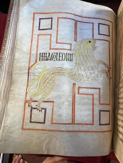 An illuminated manuscript page features a colorful illustration of a lion, accompanied by decorative borders and stylized text in Latin. The lion is depicted in a dynamic pose and is embellished with intricate patterns.