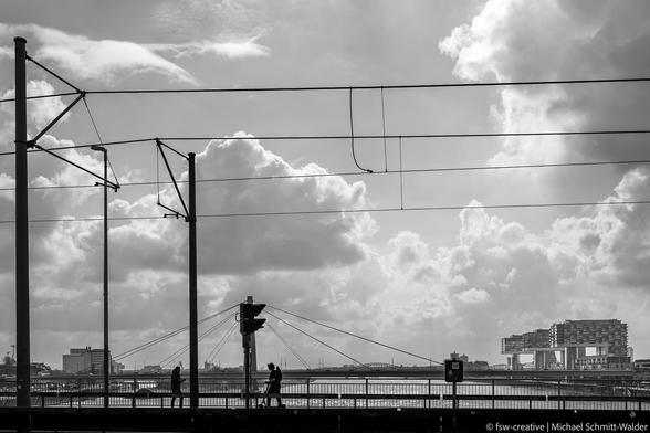 Bild in schwarz-weiss von der Deutzer Brücke in Köln mit Blick auf die Kranhäuser und südlich den Rhein entlang. Menschen mit Rädern und zu Fuss eilen zu den nächsten Terminen oder in den Feierabend. Eine ruhige und typisch kölsche Stimmung liegt in der Luft.