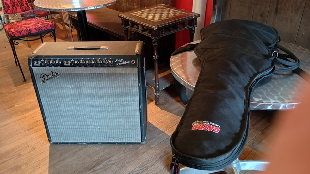 Fender Super Reberb 4x10 combo amp belonging to a local rock hero, in his shop, no longer for sale now that he's let me try it out. Probably not because of my timidity.
It's about 30 inches square, ten inches deep, with silver grille cloth and a black panel with about 14 knobs and a red jewel light.
It's on the wood floor of a restaurant at 2:30 PM next to a chess table and a couple of round brushed-steel dining tables.
The sun is shining behind it and it casts a shadow toward the camera like a monolith.
