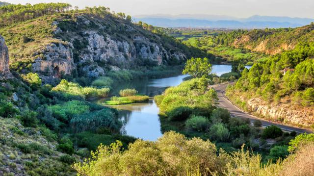 Aquesta imatge mostra un paisatge muntanyós serè amb un riu sinuós que serpenteja per una vall exuberant. L'aigua sembla tranquil·la i reflecteix el cel a la seva superfície. La vegetació és abundant, amb una barreja d'arbres i arbustos de fulla perenne que cobreixen els vessants dels turons. Un camí sinuós travessa la vall, afegint un toc de civilització al paisatge natural. A la distància, les muntanyes es poden albirar, aportant una sensació d'escala i profunditat a l'escena. La llum suau suggereix que la imatge podria haver estat presa durant l'hora daurada, potenciant els colors i creant un ambient tranquil i tranquil.