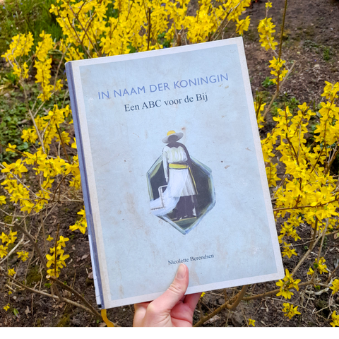 A white hand holding up a book in front of the yellow flowers of a forsythia. The cover is grey with an illustration of a woman wearing a hat. Above it the title in Dutch: In Name of the Queen, an ABC for the bee.