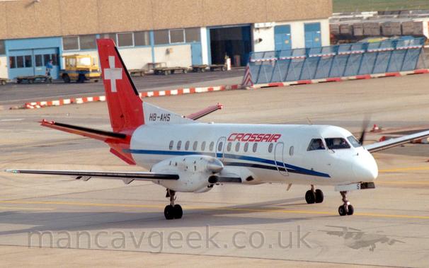 Side view of a white, twin propellor-engined airliner with a red rear-fuselage and tail, taxiing from left to right.
There is a black and blue stripe running along the body from the nose, under the passenger cabin windows, as well as red "CrossAir" titles on the upper forward fuselage over the windows.
There is a large white cross on the red tail.
Spinning propellors on the engines are blurred into barely visible smears.
Grey concrete apron fills the foreground and most of the background, with a brown and white building stretching across the frame.