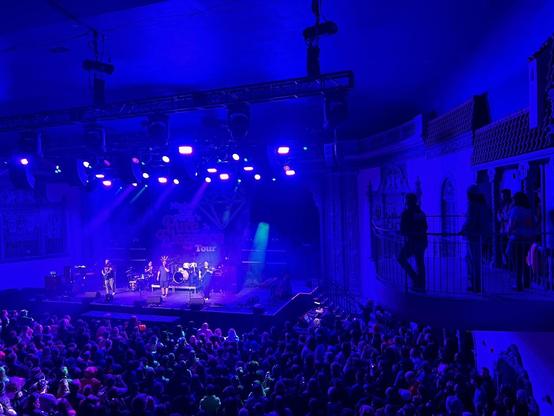 Picture of a crowded theater lit with purple and blue lights, seen from above & behind. The band Psychostick performs on stage. On a small balcony to the right, several people (including Brian & Danny of NSP) look over the scene.
