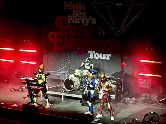 Picture of the band TWRP performing on stage, lit by white & neon red lights. Each wears a bright, colorful costume. Doctor Sung plays keyboard & sings, Havve Hogan plays drums in the back, and Commander Meouch & Lord Phobos play bass & guitar next to each other.