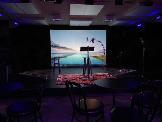 A dimly lit stage setup featuring a wooden stool, a music stand, and a desk lamp. A large screen displays a serene landscape with water and mountains. In the foreground, round tables with black chairs are arranged.
