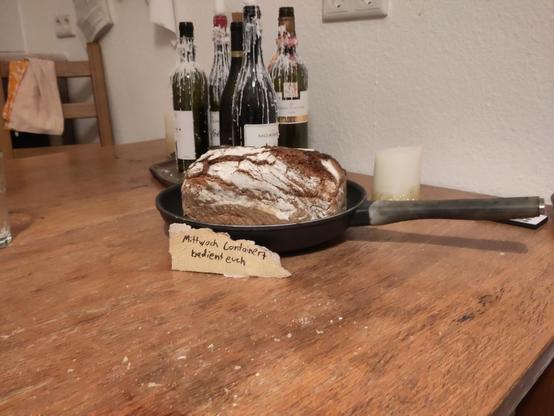 A table with a pan on it, in which is a loaf of bread
a carbord sign stands in front of it
On it is written "Mittwoch Containert,  bedient euch" which means "dumpster dived on wednesday, help yourselves" in German
Behind the bread on the table are candles made out of vine bottles