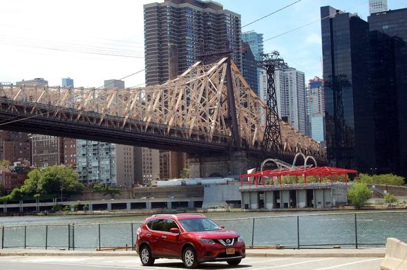 Photograph of a red car driving down the west side of Roosevelt Island with the Queensboro Bridge and Manhattan in the background.