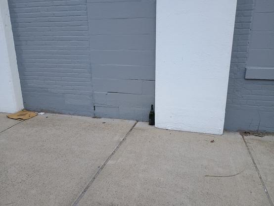A photo of a sidewalk.  There is a building with brick walls painted dark grey, and square concrete columns painted white.  Kind of a bauhaus look.  Against one of the columns is an open bottle of champagne.