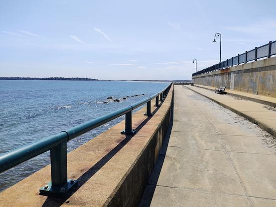 A sunny coastal walkway. To the left is the blue ocean with some rocks visible near the shore. A green railing runs along the edge of the walkway, separating it from the water. To the right, a concrete path stretches into the distance, bordered by a low seawall and a black metal fence. A few benches and lampposts are visible along the walkway under a clear, blue sky with a few wispy clouds.