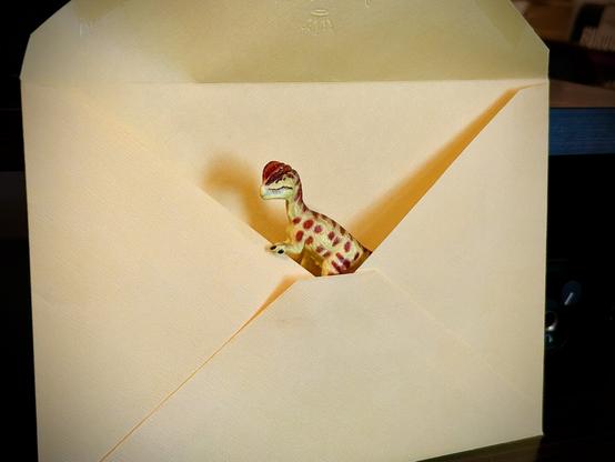 A green Dilophosaurus with brown spots sitting inside of a large, yellow envelope ready to be mailed.